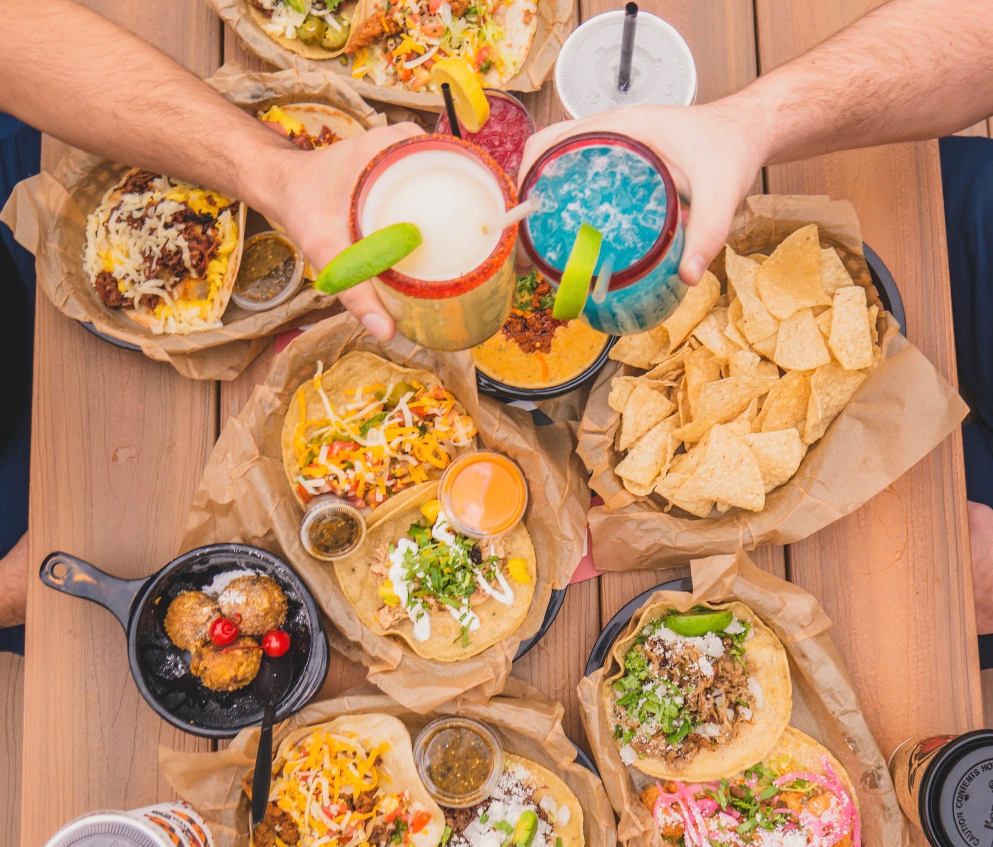 Overhead view of hands holding colorful cocktails above a table of tacos, queso and tortilla chips