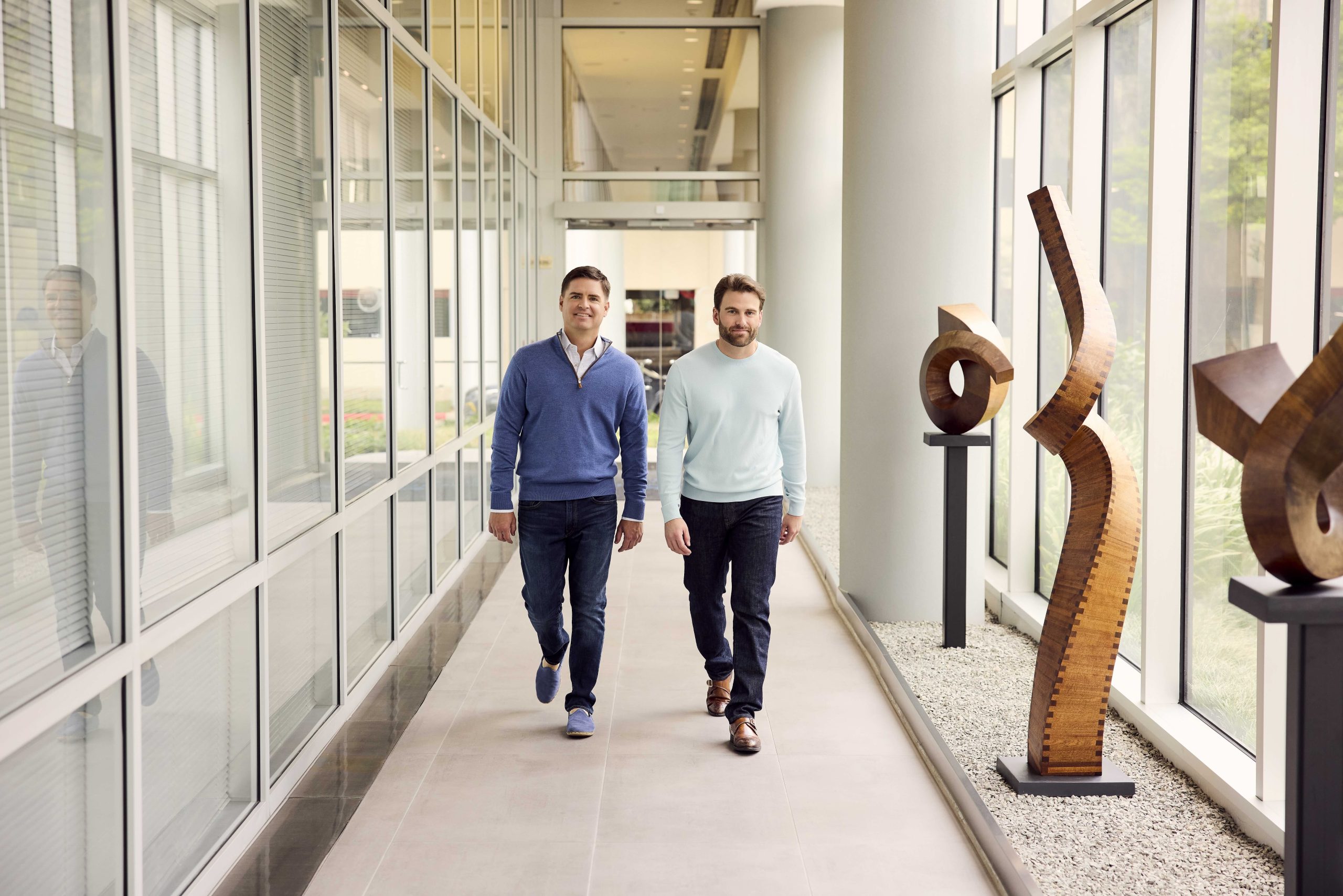 Two colleagues walking down a glass-walled corridor lined with carved wood sculptures